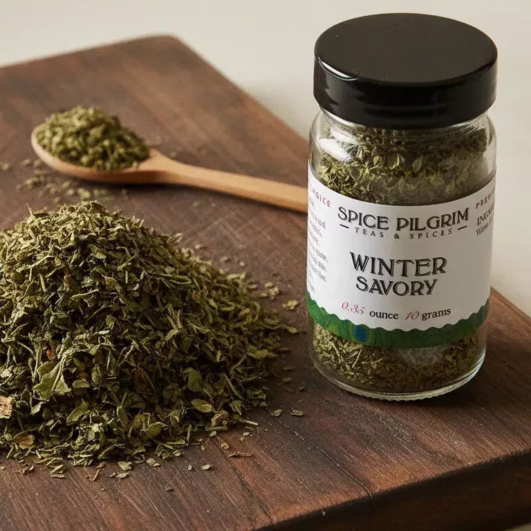 Spice Pilgrim Winter Savory dried herb jar beside a loose pile of the herb and a wooden spoon on a cutting board