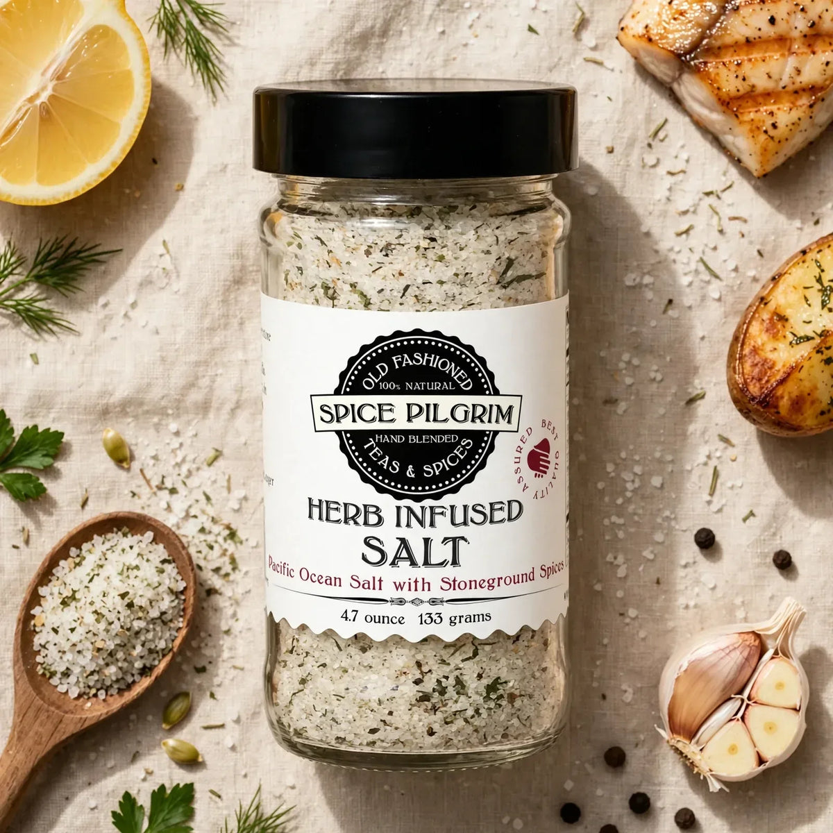 A glass jar of Herb Infused Salt sits centered on a warm linen surface, its pale green-flecked crystals catching the