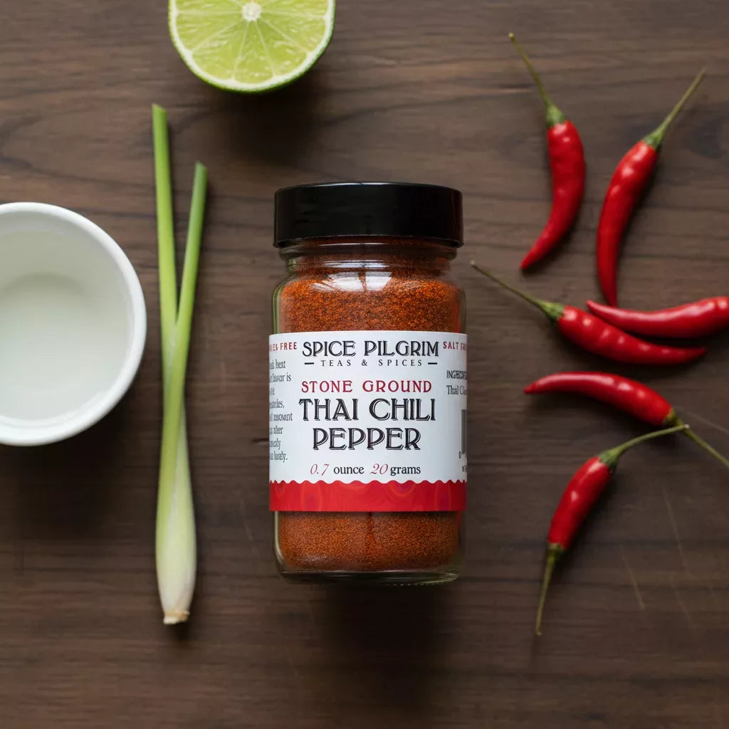 A jar of ground Thai chili sits on a dark wood surface beside five whole dried Thai chili peppers arranged in a loose