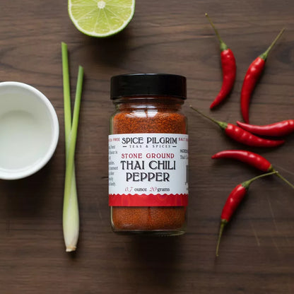 A jar of ground Thai chili sits on a dark wood surface beside five whole dried Thai chili peppers arranged in a loose