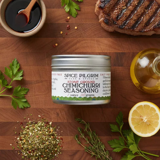 Spice Pilgrim Stone Ground Chimichurri Seasoning tin, 1 ounce, surrounded by fresh herbs, grilled steak, lemon, and olive oil on a wood cutting board