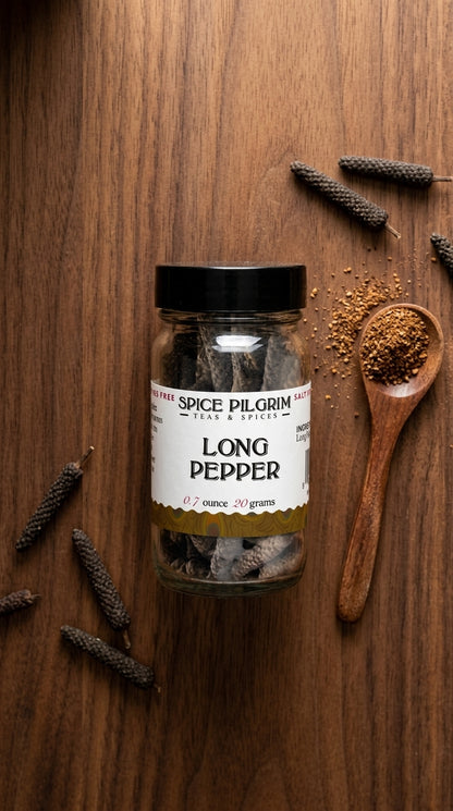 A single glass jar of long pepper sits centered on a warm walnut surface, its dark slender pods visible through clear