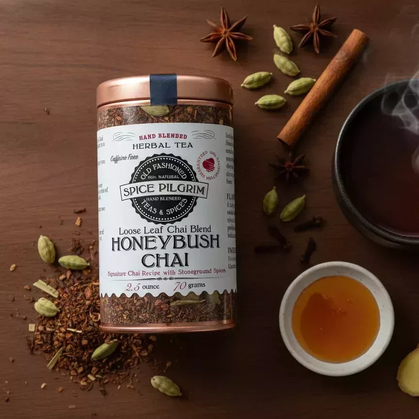 A steaming ceramic cup filled with deep amber Honeybush Chai sits slightly off-center on a dark walnut surface, wisps