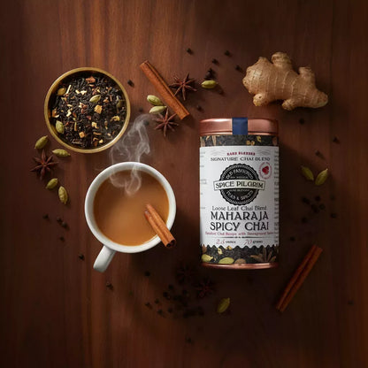 Spice Pilgrim Maharaja Spicy Chai loose leaf blend tin beside a steaming cup of chai, loose tea, and whole spices including ginger, cinnamon, star anise, and cardamom