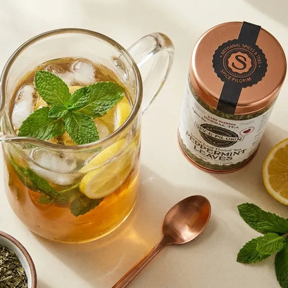 Glass pitcher of iced peppermint tea with mint leaves and lemon slices beside a Spice Pilgrim Peppermint Leaves tin, copper spoon, and lemon halves