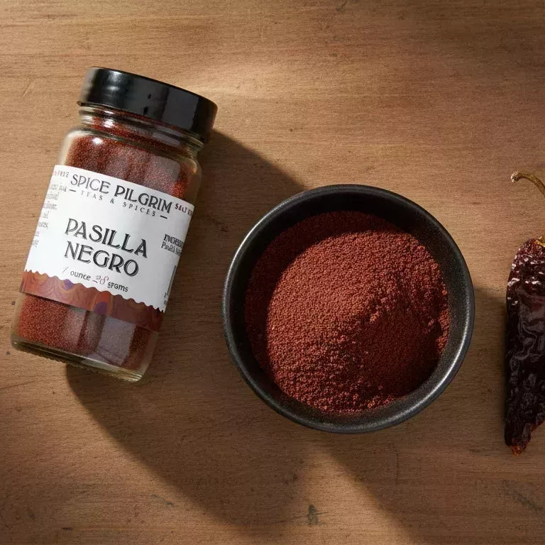 Spice Pilgrim Pasilla Negro spice jar next to a bowl of ground pasilla negro powder and a whole dried pasilla negro pepper